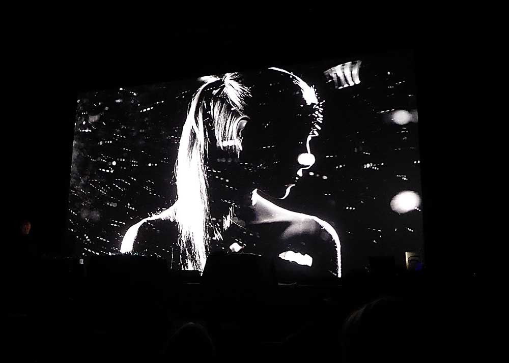 Black and white image of a woman with a ponytail on a film screen seen from the audience perspective. Woman's head is turned to the side and her face is in shadow with light shapes flying across the screen. On the left of the screen stands Scanner with his laptop performing a live film score