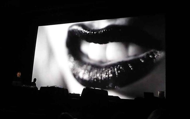 Black and white image of a close-up of a woman's mouth wearing lipstick, displaying her teeth, on a cinema screen. On the left of the screen stands Scanner with his laptop performing a live film score.