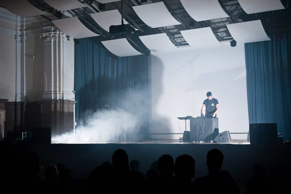 Man on stage (Haxan Cloak) seen from the audience standing over a table with black cloth and music equipment. Stage is brightly lit with smoke.