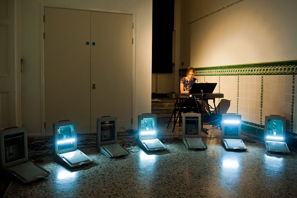 Installation by Computer Junk Orchestra of a row of computer scanners lit up. In the back of the space sits a woman with a keyboard and sheet music.