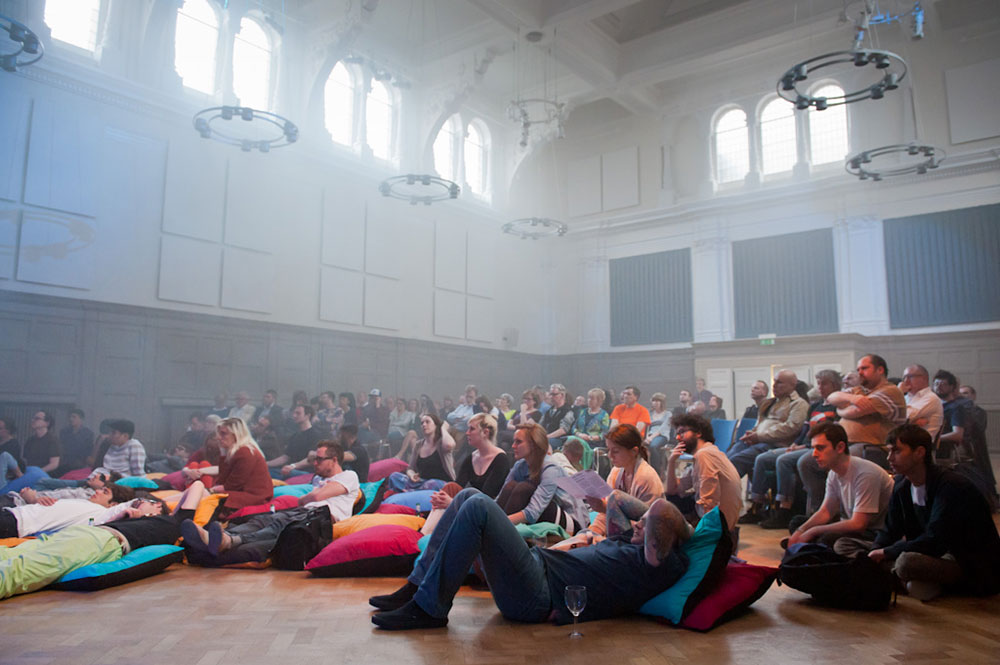 Colourful image of audience seated and lying down on bean bags on the floor of a large hall
